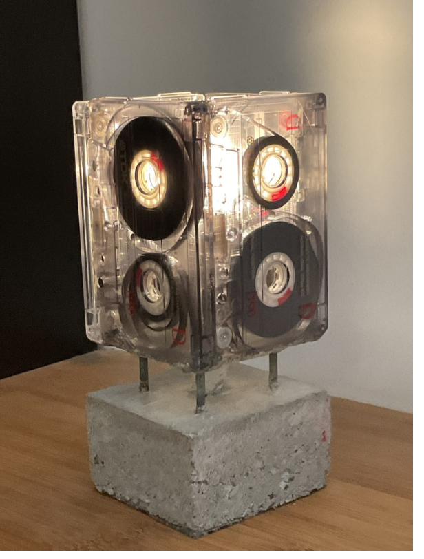 LumiK7 : lampe de chevet où la lumière traverse des cassettes audio translucides, posée sur un pied en béton